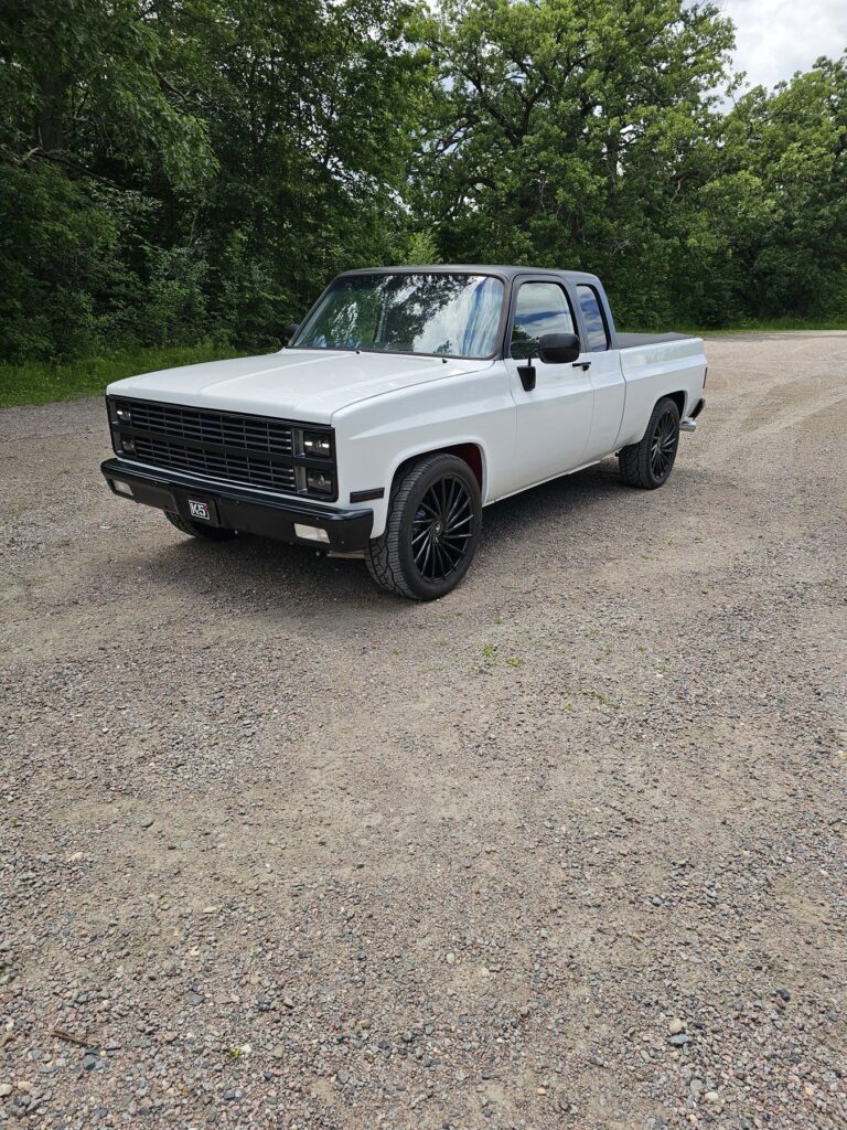 White 85 Chevy extended cab on a Frame-Werx chassis, front three-quarter on a gravel road